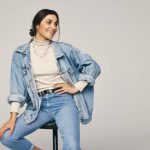 Rocking denim wear. Fashionable young woman sitting on a chair against a grey background. Happy young woman looking away with a smile on her face in a modern studio.